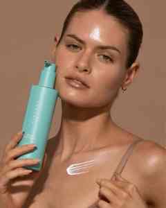 Woman holding a aqua bottle of sunscreen against a brown background.