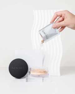 A hand pours powder from a sleek ADORN container onto a compact for flawless makeup.