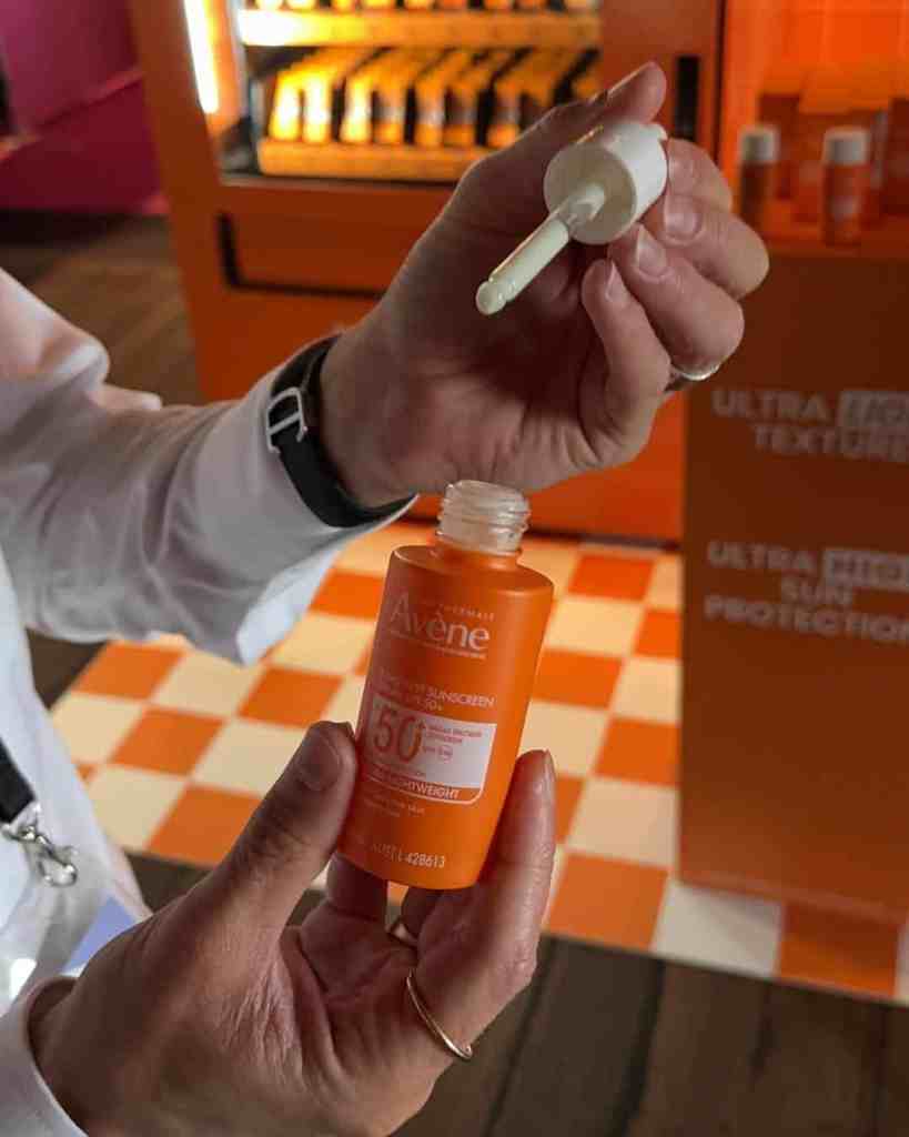 A person applying Avene SPF 50 sunscreen with a dropper in a vibrant retail setting.