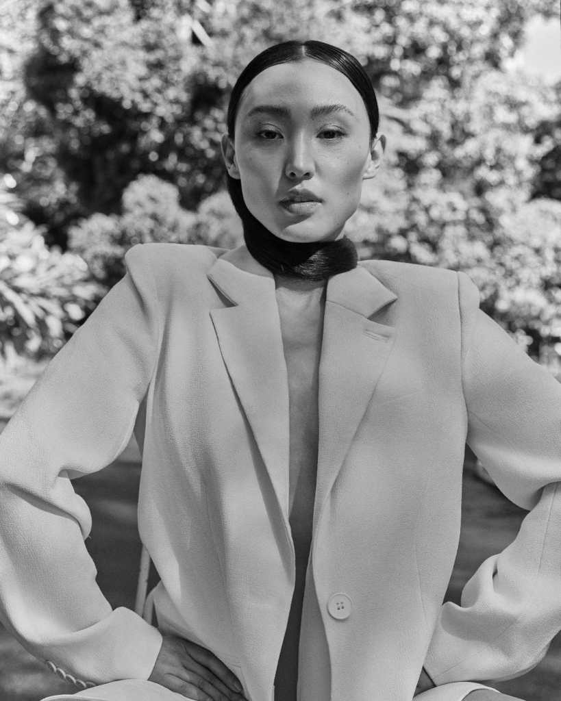 Close-up of a woman with flawless skin and sleek black hair, in a lush outdoor environment.