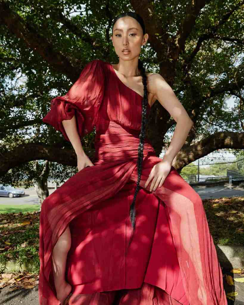 Fashionable woman in a red flowing dress posing outdoors under a tree.