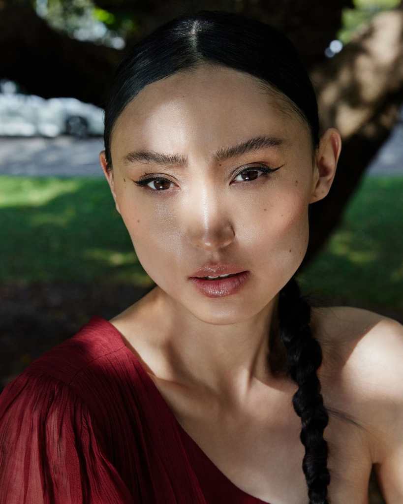Close-up of a young woman with braided hair, natural makeup, and a red top outdoors.