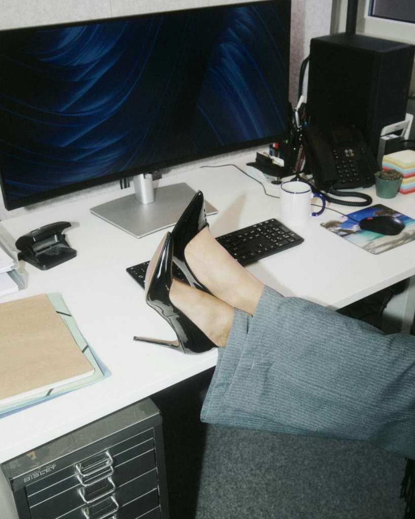 Modern office workspace with a person’s feet in high heels on a cluttered desk, featuring tech and stationery.